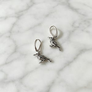 May include: A pair of silver-toned leverback earrings featuring a detailed bird charm. Each earring displays a small, sculpted bird with oxidized recessed lines to highlight the wing and feather texture, suspended from a simple polished metal hoop.