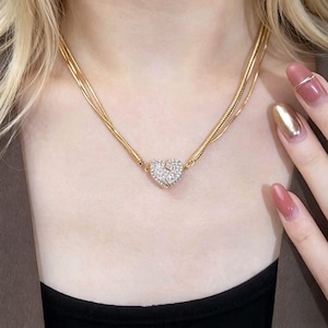May include: A gold-toned double-strand snake chain necklace featuring a central heart-shaped pendant. The heart is divided into two pieces and is encrusted with small, clear, sparkling stones that cover the entire surface of the pendant.