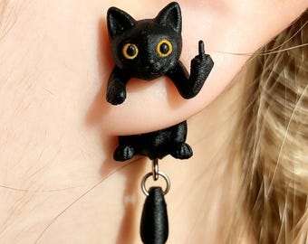 Pode incluir: Um brinco de gato preto com uma figura felina texturizada e olhos amarelos brilhantes. O gato está posicionado com as patas dianteiras sobre o lóbulo, uma pata levantada num gesto de dedo médio e um pequeno pendente de lágrima preta pendurado.