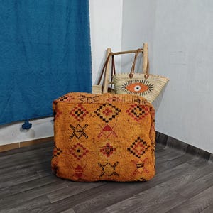 Könnte beinhalten: Ein quadratischer, orangefarbener Bodenpouf mit einem sich wiederholenden Muster aus schwarzen und roten geometrischen Symbolen. Hinter dem Pouf steht eine geflochtene Strohtasche mit einem großen, gestickten Augenmotiv in Orange, Schwarz und Weiß sowie braunen Ledergriffen.