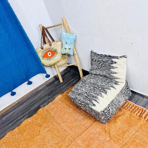 May include: A woven straw tote bag with an orange and black eye graphic, a light blue pouch, a wooden ladder, a black and white patterned floor cushion, and a large orange rug with a white diamond pattern and fringed edges.