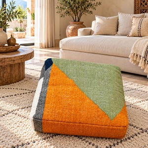 May include: A floor cushion featuring a geometric pattern with large blocks of orange, sage green, dark gray, and white. The textile has a visible woven texture and a wedge-like shape, with a flat base and an angled top surface for support.