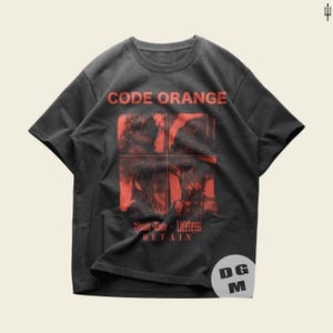May include: A charcoal gray short-sleeve t-shirt featuring the text CODE ORANGE in red at the top. Below, a red graphic is divided into four quadrants. The text Youth Code, Lifeless, and DETAIN appears in red at the bottom of the graphic.