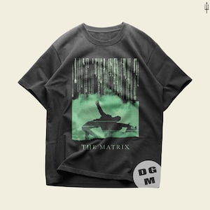 Peut inclure: Un t-shirt gris anthracite à manches courtes avec un motif vert représentant une silhouette esquivant des balles sous une pluie numérique. Le texte THE MATRIX est imprimé sous la figure. Un logo blanc circulaire avec les lettres DGM est présent.