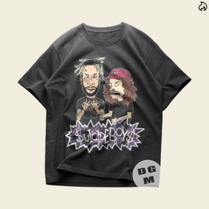 May include: A charcoal gray short-sleeve t-shirt featuring a cartoon graphic of two individuals with long hair and a red cap. Below the figures is a jagged purple lightning-themed graphic containing the text $UICIDEBOY$ in a stylized font.