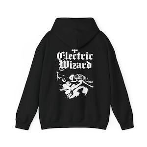 May include: A black hooded sweatshirt featuring a white graphic print on the back. The design includes the text Electric Wizard in a stylized font above an illustration of a figure with long hair and a skull, rendered in high-contrast white ink.
