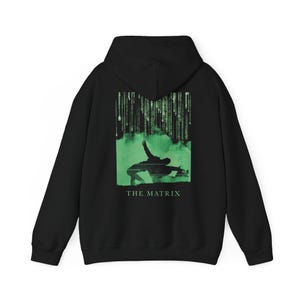 Op de afbeelding: Een zwarte hoodie met een afbeelding op de achterkant. Het ontwerp toont het groene silhouet van een persoon die achterover leunt tegen een achtergrond van vallende digitale code, met de tekst THE MATRIX in het groen onder de figuur.