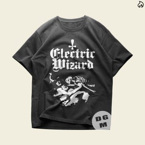 May include: A black short-sleeve t-shirt featuring the white text Electric Wizard in a gothic font with an inverted cross symbol above it. Below the text is a white graphic illustration of a skeleton and a figure. A circular DG M logo is visible.