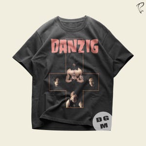 May include: A black short-sleeve t-shirt featuring the word DANZIG in pink letters at the top. Below the text is a large orange cross outline containing photographic portraits of four band members arranged in a cross formation on the chest.