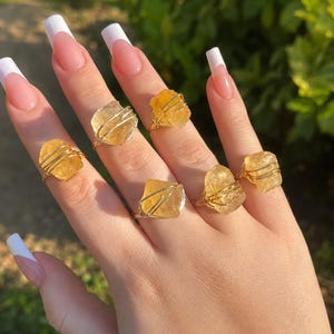 May include: Five rings featuring raw, translucent yellow citrine crystals secured with thin gold-toned wire wraps. Each ring showcases a unique, jagged gemstone set on a simple metallic band, highlighting the natural texture and golden hue of the stones.