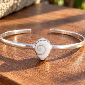 Op de afbeelding: Een zilveren armband met een centrale druppelvormige witte Shiva-oogschelp met een natuurlijk bruin spiraalpatroon. De gepolijste metalen band is aan de achterkant open en heeft een gladde, reflecterende afwerking die het detail benadrukt.