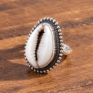 May include: A ring featuring a white cowrie shell set in a silver-toned metal frame. The oval-shaped shell is surrounded by a decorative border of small, raised silver beads. The metal band is smooth and polished, extending from the central setting.