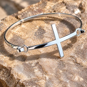 Może przedstawiać: Polerowana bransoletka typu bangle w kolorze srebrnym z centralnie umieszczonym krzyżykiem. Bransoletka składa się z cienkiej, okrągłej obręczy połączonej z krzyżem w dwóch punktach za pomocą małych, zintegrowanych oczek, tworząc gładki, lśniący wzór.