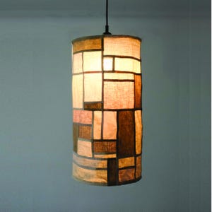 May include: A cylindrical pendant lamp featuring a patchwork design of rectangular fabric panels in various shades of beige, tan, and brown. The light emits a warm glow through the textured material, which is suspended from a single black cord.