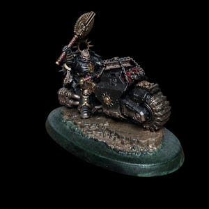 May include: A miniature figurine of a warrior with a skull-like helmet riding a black armored motorcycle. The warrior holds a gold-colored staff with a winged emblem aloft. The motorcycle features metallic silver details and chains.