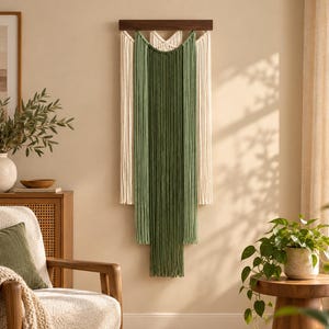 May include: A wall hanging featuring long, vertical cotton cords in cream and sage green. The cords are suspended from a horizontal dark wood bar, arranged in layered sections with the green cords centred over the cream-coloured base layer.