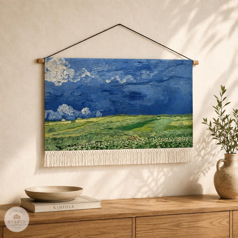 May include: A rectangular wall hanging featuring a printed landscape painting of a green field under a dark blue, cloudy sky. The textile piece is suspended from a thin black cord attached to a wooden rod and finished with a white fringe at the bottom.