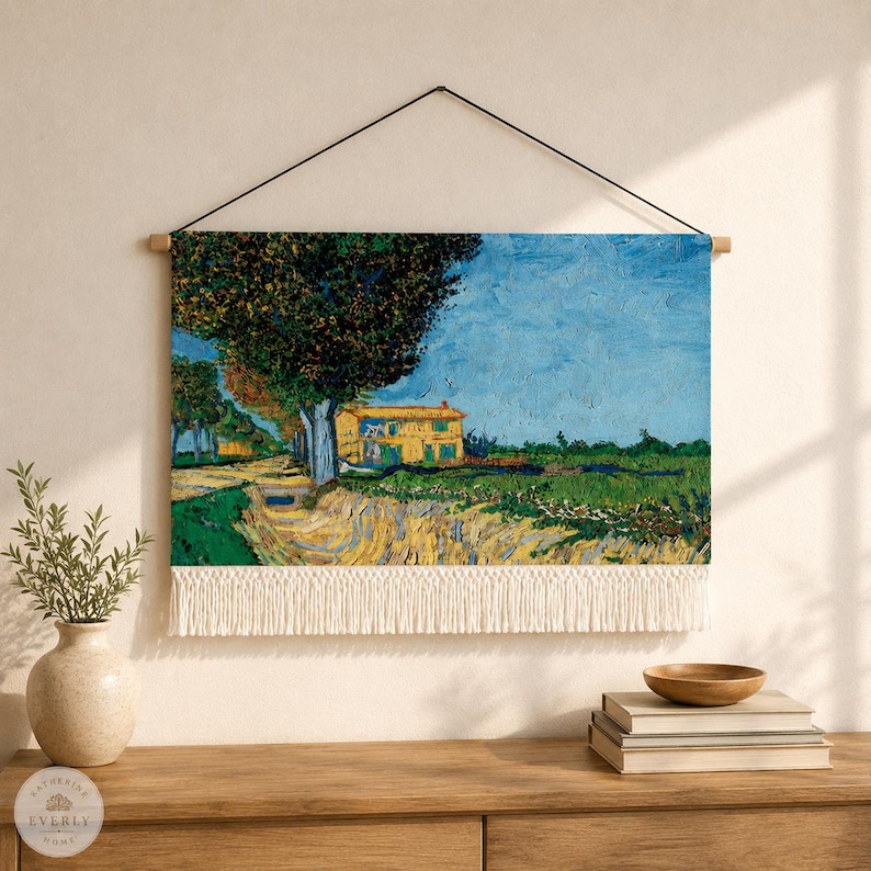 May include: A rectangular wall hanging featuring a reproduction of a landscape painting with a yellow house, a large dark tree, and a blue sky. The fabric print is suspended from a wooden rod with a black cord and has a white fringe at the bottom.