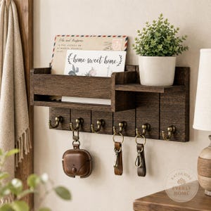 May include: A dark brown wooden wall-mounted organiser featuring a mail slot, a small shelf holding a potted green plant, and six brass-toned hooks. The mail slot contains an envelope with the text home sweet home and other printed details.