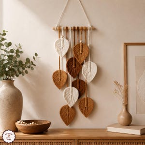 Op de afbeelding: Een macramé wandhanger met een horizontale houten stok met hangende koorden en meerdere bladvormige vezelelementen in tinten crème, beige en bruin. Elk blad is in het midden aan de bovenkant versierd met een kleine houten kraal.