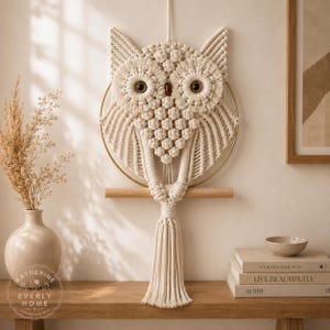 Puede incluir: Un tapiz de pared de macramé color crema con forma de búho, que presenta intrincados patrones de nudos, dos ojos circulares de color marrón oscuro y una larga borla central. El búho está montado sobre un aro metálico dorado y un listón.