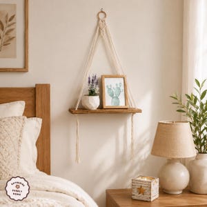 May include: A rectangular wooden wall shelf suspended by cream-coloured macrame cord with a knotted design. The shelf holds a white geometric planter with purple flowers and a framed print of a green cactus. The shelf hangs from a circular ring.