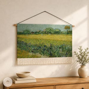 May include: A rectangular wall hanging featuring a reproduction of a landscape painting with a yellow field, green trees, and blue irises. The fabric print is suspended from a wooden rod with a black cord and finished with a cream fringe at the bottom.