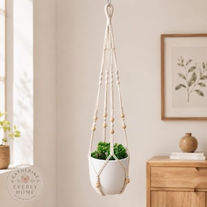May include: A cream-coloured macrame plant hanger featuring intricate knotted patterns and smooth, round wooden beads. The hanger supports a white ceramic pot containing a small green plant, suspended by a single loop at the top.