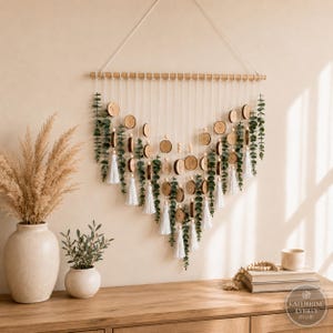 Peut inclure: Une décoration murale composée d'une tige en bois horizontale avec des cordons blancs suspendus en forme de V. Les cordons sont ornés de petites tranches de bois rondes, de feuilles d'eucalyptus vertes et de pompons blancs.