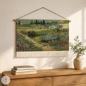 May include: A rectangular wall hanging featuring a printed landscape painting with a path, trees, and colorful vegetation. The fabric piece is suspended from a wooden rod by a black cord and finished with a long, white fringe along the bottom edge.