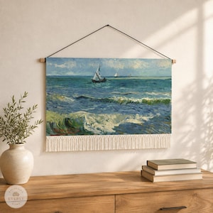 May include: A rectangular wall hanging featuring a seascape painting of a sailboat on blue ocean waves under a cloudy sky. The canvas is suspended from a wooden rod by a black string and finished with a cream-colored fringe along the bottom edge.