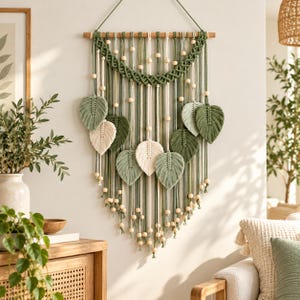 May include: A macrame wall hanging featuring a wooden dowel, sage green and cream cotton cords, and natural wooden beads. The design includes multiple leaf-shaped macrame elements in varying shades of green and cream, arranged in a layered pattern.