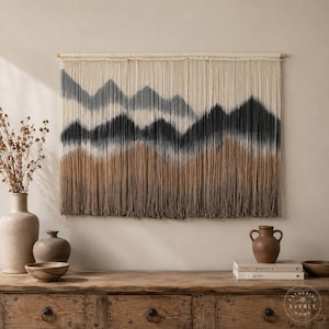 May include: A rectangular macrame wall hanging featuring a mountain landscape design in shades of cream, grey, and brown. The piece is suspended from a wooden dowel and consists of numerous vertical cotton cords with a dip-dyed gradient effect.