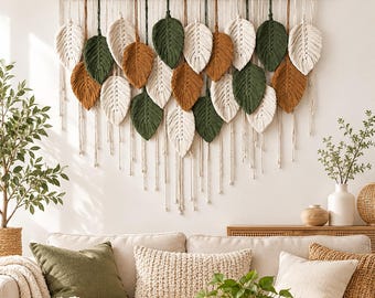 Puede incluir: Un tapiz de macramé que presenta numerosos elementos de fibra en forma de hoja en tonos crema, verde oliva y terracota. Las hojas están suspendidas de una varilla de madera horizontal mediante cordones individuales, creando un diseño texturizado.