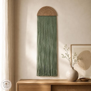 May include: A wall hanging featuring a semi-circular top section made of woven tan fibres, from which a long, dense fringe of sage green cord hangs vertically. The piece is structured with a flat, arched upper border and straight, flowing strands.