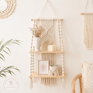 May include: A two-tiered wooden wall shelf suspended by cream-coloured macrame cord with intricate knotting patterns. The shelves hold a white face-shaped vase with dried flowers, a square bubble candle, a framed print, and a glass jar with a macrame cover.