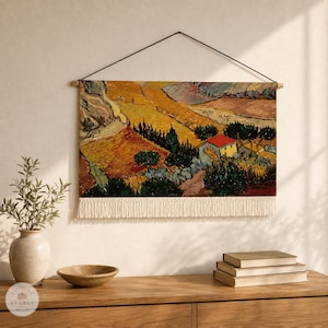 May include: A rectangular wall hanging featuring a landscape painting with golden fields, dark green trees, and a small house with a red roof. The artwork is suspended from a wooden rod by a black cord and finished with a cream-colored fringe at the bottom.