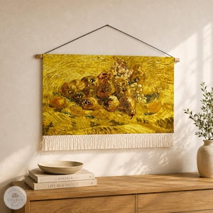 May include: A rectangular wall hanging featuring a reproduction of a still life painting with yellow tones, depicting a cluster of fruits including grapes and pears. The textile piece is suspended by a black cord and features white fringe at the bottom.