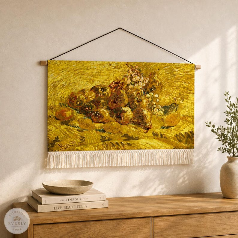 May include: A rectangular wall hanging featuring a reproduction of a still life painting with yellow tones, depicting a cluster of fruits including grapes and pears. The textile piece is suspended by a black cord and features white fringe at the bottom.