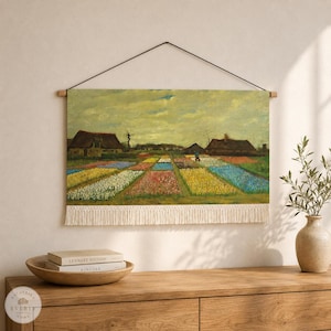 May include: A rectangular wall hanging featuring a printed landscape painting of a flower field with rows of colorful blooms, two rustic houses, and a cloudy sky. The textile piece is suspended by a thin black cord and has white fringe.