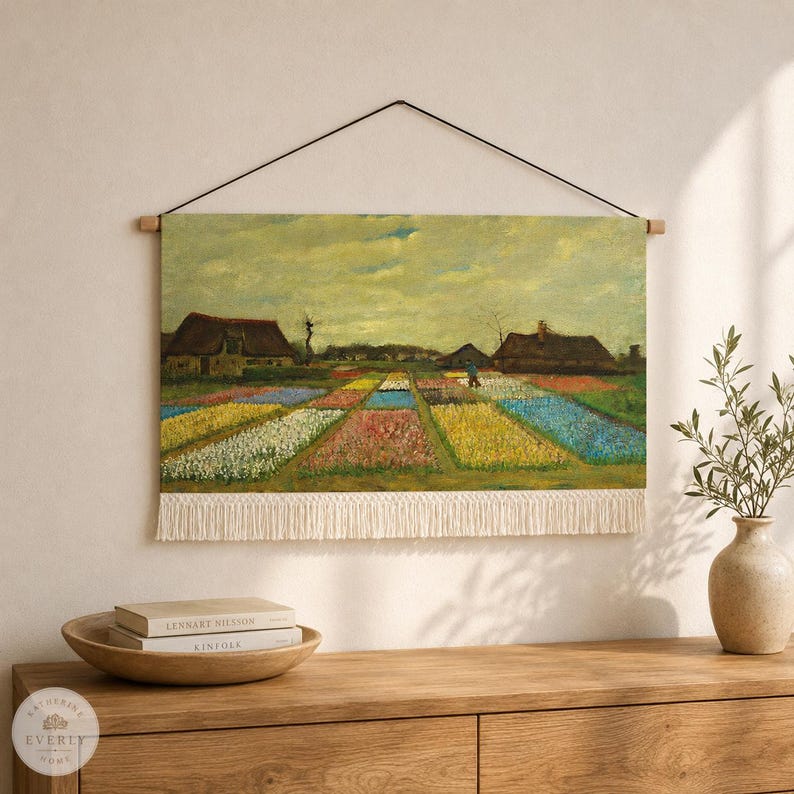 May include: A rectangular wall hanging featuring a printed landscape painting of a flower field with rows of colorful blooms, two rustic houses, and a cloudy sky. The textile piece is suspended by a thin black cord and has white fringe.