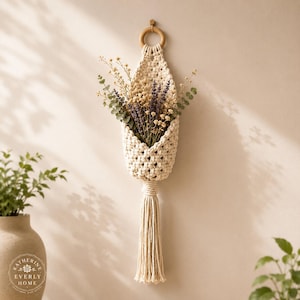 May include: A cream-coloured macrame wall hanging featuring a woven pocket suspended from a wooden ring. The pocket holds a bouquet of dried lavender, yellow flowers, and green eucalyptus stems. A long, thick macrame tassel hangs from the bottom.
