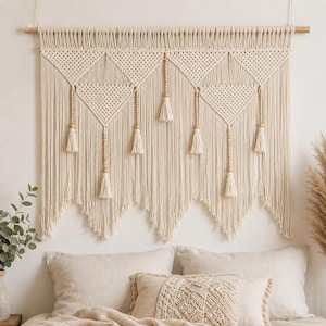 May include: A large off-white macrame wall hanging suspended from a wooden rod. The design features intricate knotted diamond patterns, vertical rows of light wooden beads, and long fringe tassels hanging from the bottom of each geometric section.