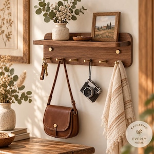 May include: A wall-mounted dark wood shelf featuring a flat top surface and six brass-colored hooks. The shelf holds a small white speckled vase with greenery, a wooden bowl, and a framed landscape print. Items hanging include a brown leather bag, a camera, and a striped beige towel.