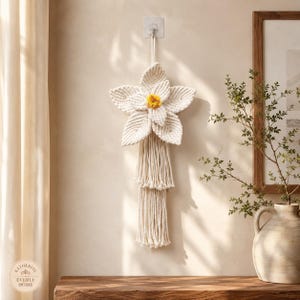 May include: A wall hanging featuring a cream-coloured macrame flower with five large, textured petals and a bright yellow centre. The flower is suspended by a cord and includes two layers of long, vertical fringe tassels hanging from the base.