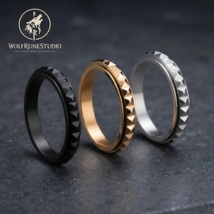 May include: Three rings arranged in a row, featuring a black ring, a gold-toned ring, and a silver-toned ring. Each band has a textured outer surface with a repeating pyramid-shaped stud pattern. The rings are displayed below the WolfRuneStudio logo.