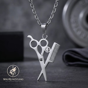May include: A silver-toned metal pendant necklace featuring a pair of hair styling scissors crossed with a hair comb. The pendant includes a small, clear, round embedded crystal accent. The piece hangs from a matching silver-toned metal chain.