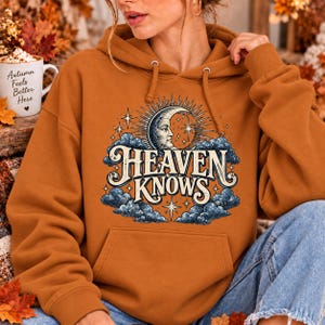 May include: A burnt orange hooded sweatshirt featuring a graphic print of a crescent moon with a face, surrounded by stars and dark blue clouds. The text "HEAVEN KNOWS" is printed in a stylized cream font across the center of the design.