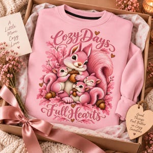 May include: A light pink crewneck sweatshirt featuring a graphic of three squirrels holding acorns surrounded by pink leaves. The text reads Cozy Days Full Hearts. The sweatshirt is packaged in a gift box with a pink ribbon and a small card.