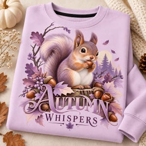 May include: A light purple sweatshirt featuring a graphic of a squirrel sitting on a tree stump holding an acorn. The design includes purple oak leaves, berries, and a forest background with the text Autumn Whispers in a stylized purple font.